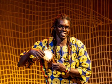 Man with dreadlocks wearing glasses and a colorful patterned shirt playing a small handheld drum on a stage with a textured gold backdrop.
