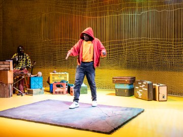 A South Sudanese man in a red hoodie and orange shirt dances while South Senegalese man plays percussion instruments in the background, set on a bright yellow stage with props like crates and suitcases