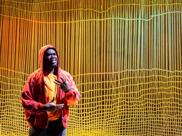 A South Sudanese man wearing a red hooded sweatshirt and orange shirt gestures with his hands, speaking on a stage with a warm-toned abstract backdrop of lines and grids.