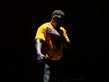 A South Sudanese man in an orange t-shirt, hand on chest, lit dramatically against a dark background, suggestive of performance or emotion.