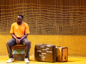 A South Sudanese man in an orange shirt and jeans sits on vintage suitcases, looking contemplative, against a backdrop of a yellow grid pattern and a brown wall, with a suitcase labeled with African city names.