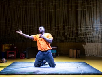 A South Sudanese man in orange t-shirt and jeans kneels on a blue stage, one arm outstretched, hand over his chest, face turned up with a look of emotion, in front of a backdrop of hanging cords and stacked luggage.