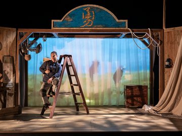 A young Chinese man sitting on a wooden ladder on a stage.