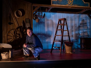 A young Chinese man squatting on a wooden stage with a ladder next to him..