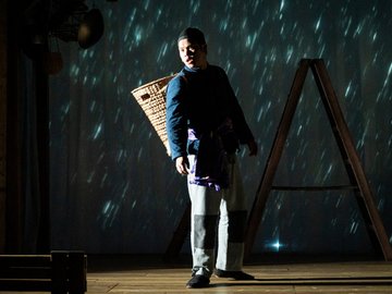 A young Chinese man standing on a stage with a basket hanging from his back, and theatrical rain behind him.