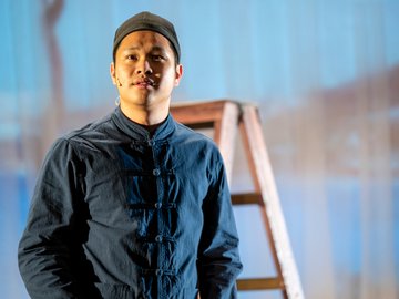 A midshot of a young Chinese man on a stage, wearing a navy shirt and hat.