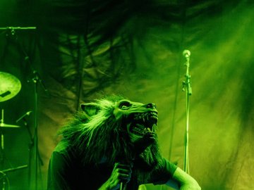 A person wearing a wolf mask kneels on stage, operating a sound mixer. There is green stage lighting.
