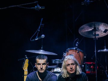 Two people sit on the edge of a stage, surrounded by rock instruments, looking direct to camera.