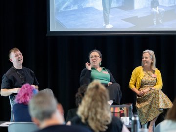 Panel discussion with three speakers on stage, audience in foreground, presentation screen behind.