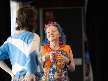 Woman with colorful hair and flamingo earrings holding a mug, talking with another person.