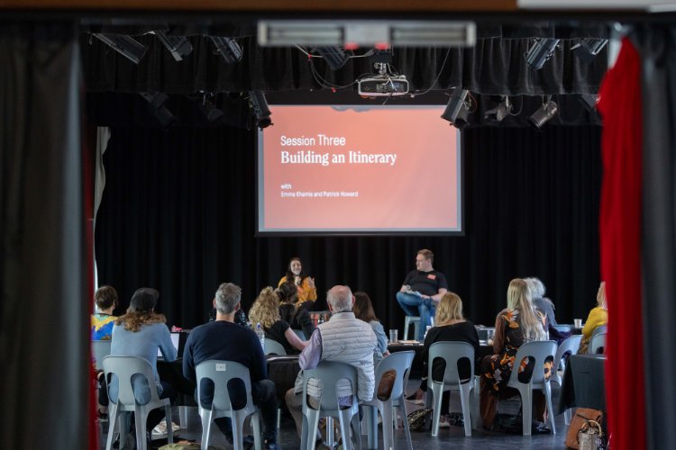 Audience watching "Building an Itinerary" stage presentation in a darkened auditorium.