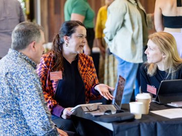 Three professionals networking with laptops at an event table, discussing business in a bright setting.