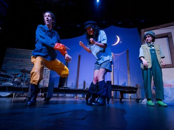 Three actors in costume on a stage set. One man in blue jacket, yellow pants, and boots gestures emphatically, another pulls up her pants, and a third in overalls looks on. A backdrop depicts a night scene with a moon.