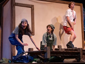 Three actors on stage with surprised expressions; the actor on the left wears a striped shirt, blue overalls, and cap, while the actor in the middle wears a green outfit and hat; the actor on the right wears a white shirt, red shorts, and holds clipboard.
