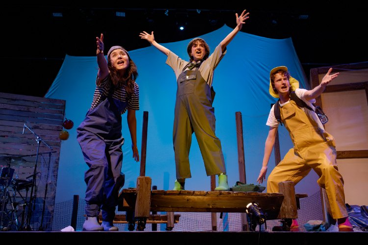 Three actors in costume on a stage, arms raised in excited gestures, appear to be performing in a play, wearing overalls and wellington boots.