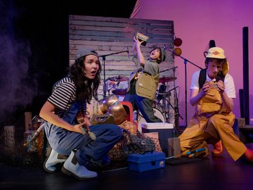 Three actors on a stage set for a play, dressed in fishing attire, one crouching with a small tool, another holding up a container excitedly, and the third seated, looking at something in their hands, all near fishing props like nets and toolboxes.