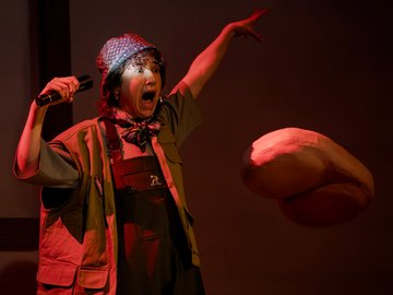 A performer on stage in a green safari-style jacket, bib overalls, and a patterned cap, holding a flashlight in one hand and with a dramatic, wide-mouthed expression and raised arm, facing a large, stylised, floating, red-lit shape.