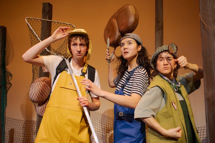 Three actors in costume, each looking upwards with surprised or concerned expressions. One holds a fishing net and wears yellow overalls, another wears a striped shirt, and the third uses a magnifying glass, dressed in a vest.