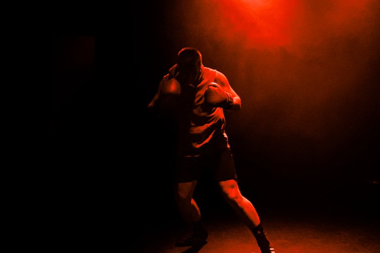 A boxer with gloved fists raised stands in a fighting stance, illuminated by dramatic red stage lighting against a dark background.