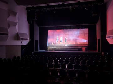 A stage with three performers and a large wall with red and white projection, viewed from a dark auditorium with rows of empty seats.
