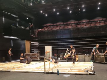 A group of theatre crew are setting up a stage wall/flat in an empty theatre with tiered seating.