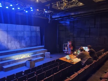 Theatre stage with a concrete-like backdrop and tiered seating, with two individuals at a control desk in the foreground and a lift in the background.
