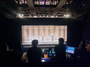 Two individuals in silhouette operate equipment in a dark control booth, facing a brightly lit stage where two performers stand before a concrete-textured backdrop.
