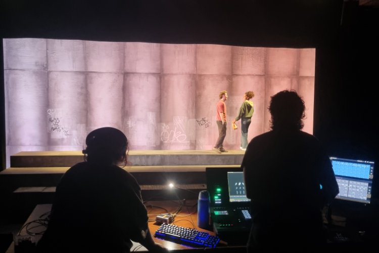 Two actors stand on a stage in front of a concrete wall backdrop with graffiti, observed from a control booth where two individuals are silhouetted by illuminated technical equipment.