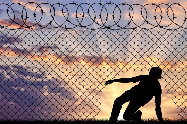 Silhouette of a person crawling through a hole in a chain-link fence, with a sunset in the background.