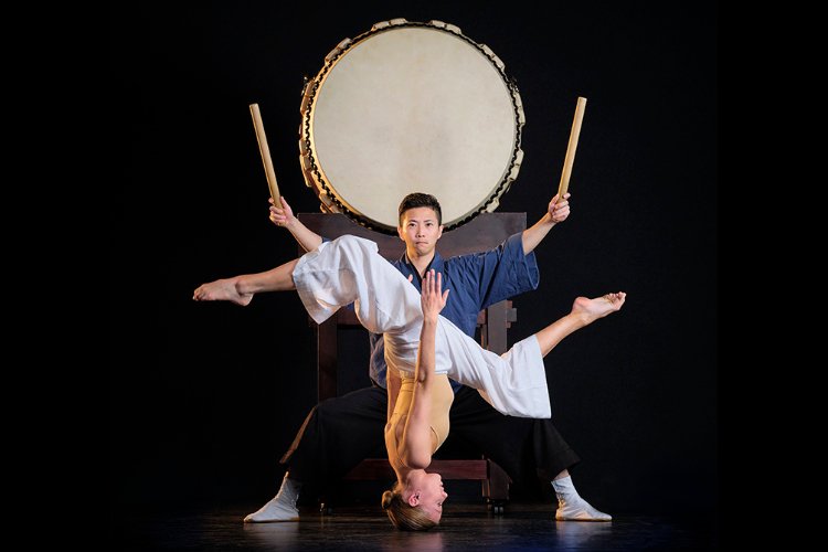 Six ballet dancers lean on a large, round drum, forming a star shape with their bodies, wearing leotards and white pants against a black background.