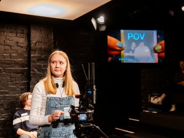 Young blonde woman in denim overalls and white turtleneck, holding a camera in a dark studio with a monitor displaying "POV", other people are seated in the background.