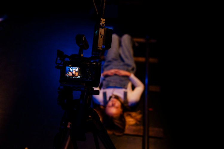 A film set, a camera on a tripod in the foreground capturing a young woman hanging upside down from a ladder in a dark, blue-lit room.