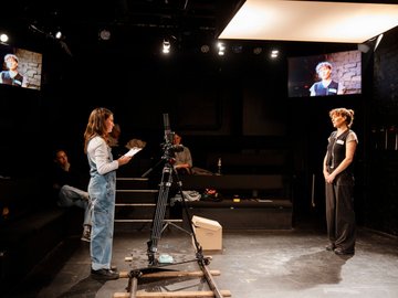 Two women in a theatre, filming a scene. One reading from a script and the other standing opposite with hands clasped. A camera is positioned in the foreground, and screens show the scene being filmed.