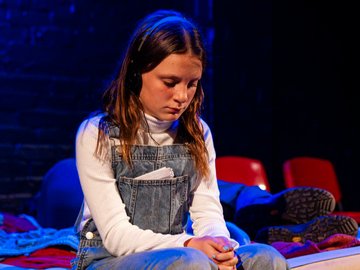 A young woman in denim overalls and a white turtleneck sweater sits with her hands clasped in her lap, looking downcast, on a bed in a theatrical setting, with a dark blue backdrop and red seats visible.