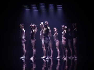 Dancers in a line with raised hands, lit by purple spotlights on a dark stage.