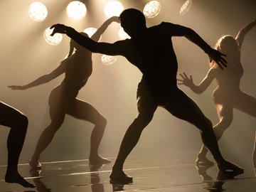 A group of silhouetted dancers performs on a stage bathed in hazy, warm lighting. Spotlights illuminate the dancers from behind, creating dramatic silhouettes and highlighting their movements.