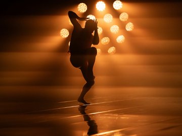 A silhouette of a dancer stands on a dimly lit stage with a backdrop of bright, warm spotlights. The dancer is slightly crouched with arms raised near his head, creating an expressive pose.