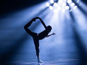 A silhouetted figure stands in a balletic balancing pose, illuminated by bright spotlights from above.