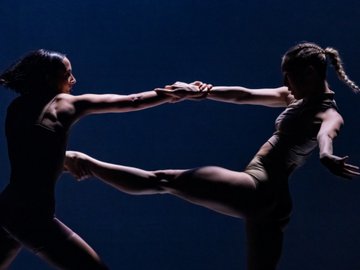 Two dancers in leotards hold hands, extending legs in a synchronized pose against a dark blue background.