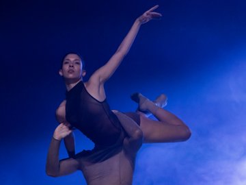 Two dancers performing a lift, one woman supported by another against a blue background with atmospheric lighting.