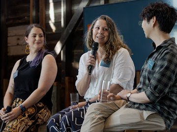 Three people seated and speaking at a panel discussion; a woman holding a microphone and speaking, another woman smiling, and a man to her right listening. They are all wearing name tags.