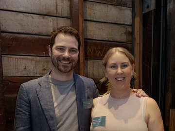 A smiling man in a blue blazer and a woman in a pink top, both wearing name tags that say "Theatre Green Book Australia", posing together in front of a rustic wooden wall.