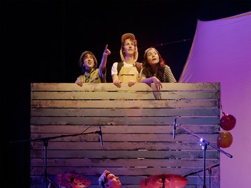 A theatrical performance with three actors looking upwards over a wooden stage wall, with a drummer in front. The actors are lit by stage lights, and the drummer sits at a Yamaha drum kit.