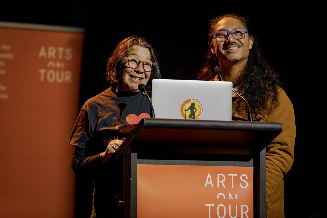 Two speakers at lectern with "Arts on Tour" branding, smiling at the audience.