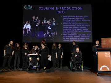 The creatives of Midnight Feast theatre company presenting on stage. The ensemble of approximately 10 people, some disabled and some non-disabled, is arranged across the stage. Some cast members are using wheelchairs. Most members are wearing