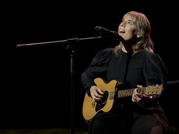 A woman with blonde hair sings and plays acoustic guitar on stage against a dark background.