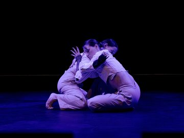 Dancers entwined in contemporary performance, kneeling with arms interlocked.