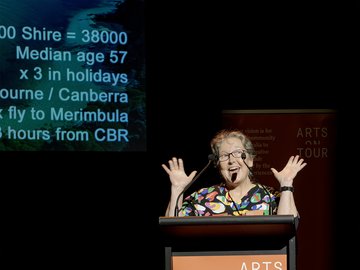 An animated speaker in a colorful shirt gestures with raised hands while standing behind a podium with "ARTS ON TOUR" displayed on it, in front of a projection screen with data.
