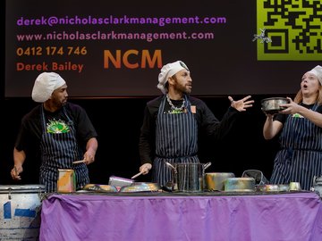 Three performers in chef hats playing culinary percussion with pots, pans, and drums on stage.
