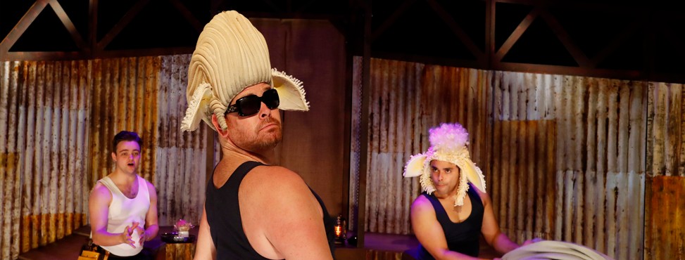 Three men in a theatrical setting. They are wearing singlets and faux fur headpieces to resemble sheep.  One is wearing black sunglasses, looking over his shoulder. The background features corrugated metal walls and dimly lit stage elements.